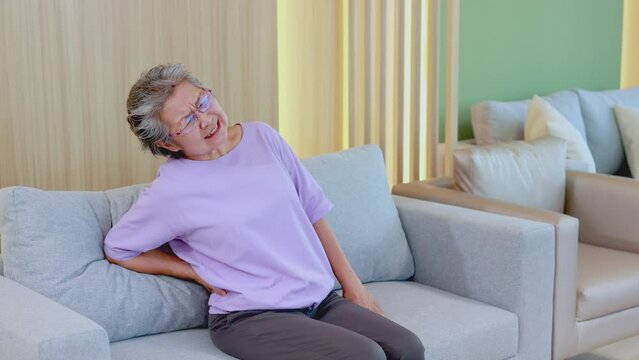 Elderly asian woman has back pain while getting up from the sofa with the help of nurse, concept of health care and treatment for the elderly.