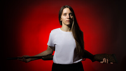 young woman with long hair in a white t-shirt holding a rifle behind her back on a red background