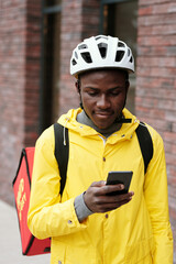 Young black man in helmet and yellow jacket looking at screen of his smartphone while delivering food to clients of online service company