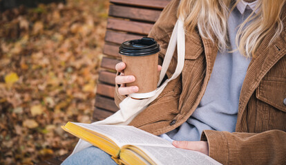 A young woman sits on a bench in an autumn park, drinks coffee and reads a book.