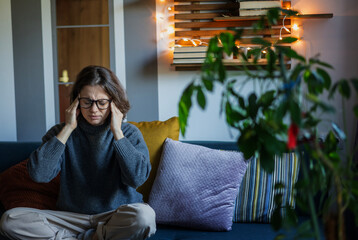 Upset young woman sitting on the couch holding on to the temporal region of the head, headaches and...