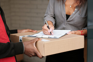 Hand of young woman with pen signing document about receiving parcel on packed cardboard box with...