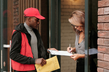 Young black man in uniform holding envelope with letter while businesswoman with pen signing...