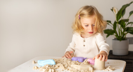 Cute happy caucasian,blonde,curly-haired toddler,baby girl playing with kinetic sand indoors.Preschool kid early development,motor,sensoric skills concept.Copy space. - obrazy, fototapety, plakaty