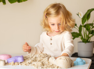 Cute happy caucasian,blonde,curly-haired toddler,baby girl playing with kinetic sand...