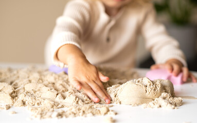 Unrecognizable cute happy caucasian,blonde,curly-haired toddler,baby girl playing with kinetic sand indoors.Child hands close-up.Preschool kid early development,motor skill, sensorics concept.