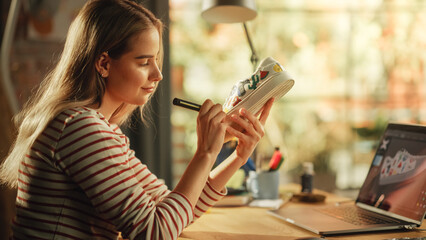Work In Creative Space: Smiling Female Artist Renovating Sneakers in front of Laptop Computer. Fashion Designer Creating Custom Made Footwear for Online Order. Post Production Concept.