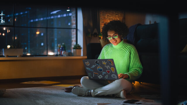 Positive Latina Female Using Laptop with Stickers at Home. Woman Resting on the Floor and Messaging on Computer with Smile on Face. Evening City View Behind the Big Window.