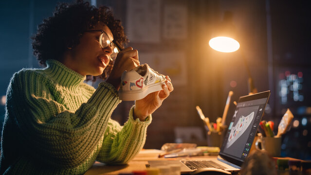 Millennial Designer Workspace: Brazilian Female Artist Sitting at the Desk and Applying her Own Designs on the Custom Shoe. Woman Working Late at Evening. People with Unique Ideas Concept.
