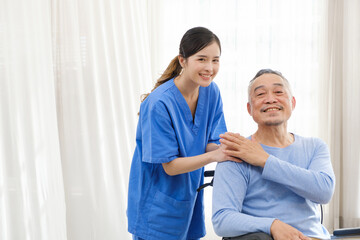 The caregiver therapist stands with an Asian senior sitting in a wheelchair and touches their hands...