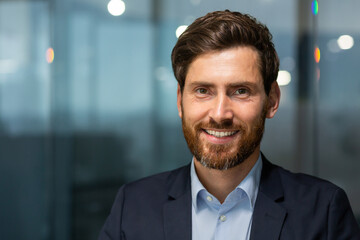 Closeup photo headshot of a mature businessman with a beard looking at the camera and smiling, a successful investor banker inside a modern office building at work, a man in a business suit evening - obrazy, fototapety, plakaty