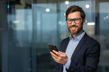 Portrait of successful mature businessman investor, man in glasses and beard smiling and looking at camera, boss in suit at work inside office holding smartphone.