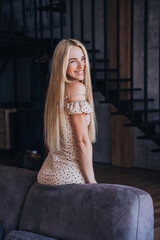 Vertical shoot of cheerful blonde young fit woman in beige dress sits on sofa at home. Gorgeous Caucasian girl with long loose hair looks at camera happily. Luxury life and beautiful people. Success.