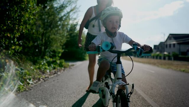 Mother Teaching her son to ride a bicycle outside in urban bike road. Successful boy wearing a helmet balancing and pedaling by himself on a bike. Handheld Slow Motion Footage