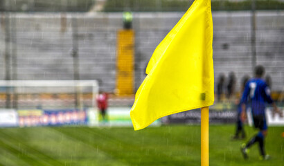 Yellow flag at one corner of football stadium and soccer corner of a soccer field with match action in the background.