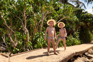 Two happy little girls in swimsuits and hat having fun on tropical country with rainforest at palm...