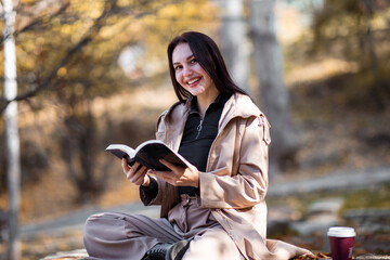 Fototapeta na wymiar Happy stylish woman portrait in autumn season with trenchcoat she is drinking coffee and reading book.
