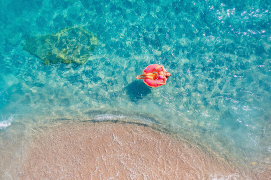 Woman swimming on pink inflatable donut in turquoise sea with wave, aerial top view