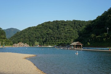 Sandy shore of water with people swimming against a green forested hills