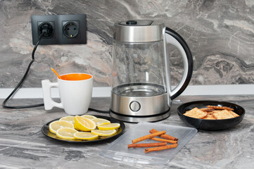 Tea drinking. Electric kettle, teacup, cookies, cinnamon and lemon on a kitchen table