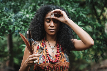 retrato de una mujer afroamericana al aire libre haciendo un ritual, con una mano sostiene algunas plumas y un rollo de hojas de salvia y con la otra coloca un cristal de cuarzo en su frente. 