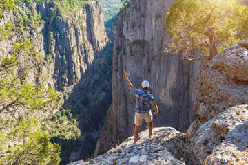 Man in checkered blue shirt and cap stands on the edge of deep canyon cliff, raising one hand up,...