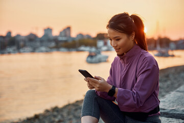Smiling Asian athletic woman texting on cell phone at sunset.
