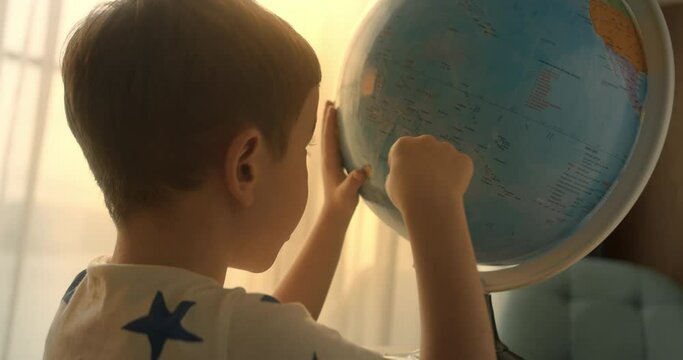 Child is studying while sitting at home in rays sun in room.Kid in morning light, Cute boy Child with Globe and magnifying glass, technology for schoolboy, child looks at globe, explores world, learns