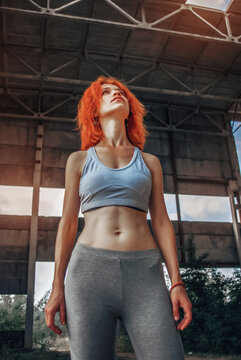 Strong female athlete wearing sportswear taking break after exercising. Fit woman standing inside abandoned warehouse after workout.