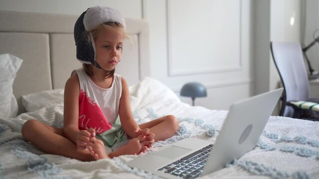 Child in funny hat watching video on laptop sitting sofa at home. Little caucasian girl enjoy cartoons on laptop sitting bed in bedroom. Joyful preschool kid girl use laptop computer