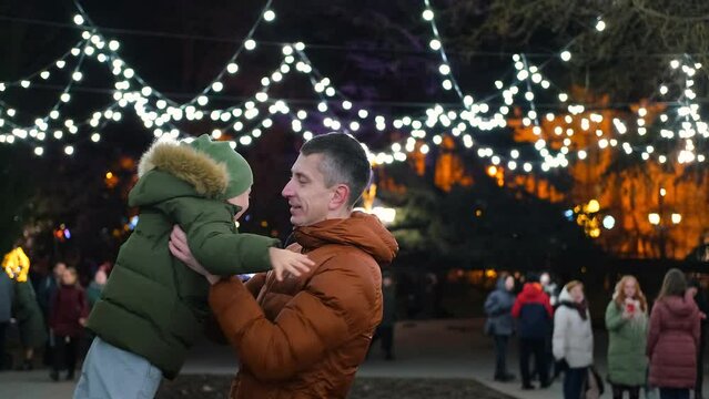 A father holds his son in his arms against the backdrop of Christmas lights. Dad turns son on the street.
