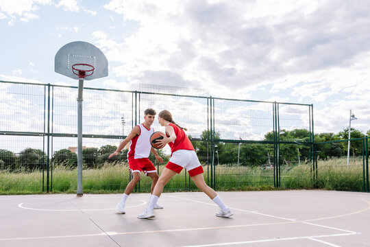 Young man and woman playing basketball on sports ground in park