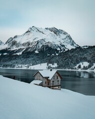 Fototapeta premium Vertical shot of a single house in front of a snowy mountain and a frozen lake