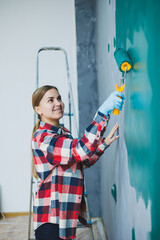 Young smiling pretty woman in shirt holding roller in right hand and painting wall. Repair in the room.