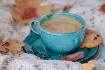 Autumnal composition with white knitted scarf, blue cup of coffee, cookies and dry yellow leaves on a table. Autumn mood.