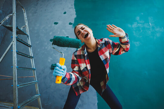 An emotional woman paints the walls. Young smiling pretty woman in shirt holding roller in right hand and singing. Repair in the room.