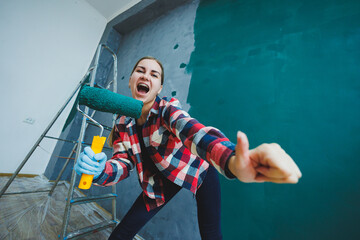 An emotional woman paints the walls. Young smiling pretty woman in shirt holding roller in right...