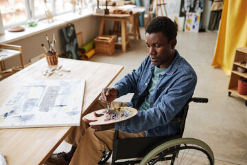 Young African American man with disability mixing paints on palette while sitting on wheelchair in studio of arts by table with canvas