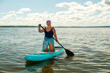 A Jewish woman in a headscarf in a pareo on her knees on a SUP board swims in the lake on a sunny day.