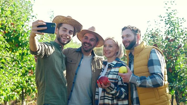 Portrait of young Caucasian cheerful happy people gardeners taking selfie photo with smartphone and posing with apples crop to camera. Outdoors. Males and female making picture with phone in orchard.