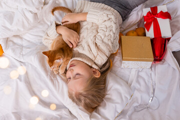 a little blonde girl in a knitted sweater is lying in bed with a red cat and gifts for the New Year, top view. Symbol of the Year, 2023