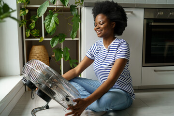 Happy smiling young black woman enjoying fresh air while cooling by electric fan at home, sitting...