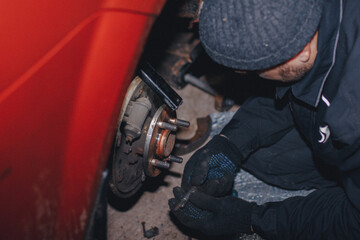 In the evening, in the garage, a mechanic in uniform and protective gloves is repairing a wheel on a car. The mechanic holds the part in his hands and examines it. Replacing worn brake discs.