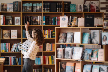 Teen girl among a pile of books. A young girl holding books with shelves in the background. She is...