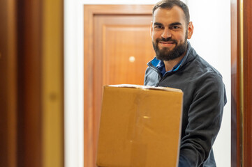 Bearded man delivering a package. Holding a parcel in front of the door. Looking at camera