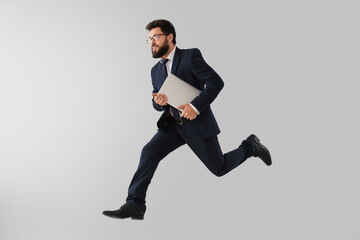 Handsome bearded businessman with laptop jumping near grey wall