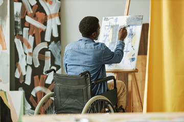 Rear view of African American male painter in wheelchair painting on canvas while sitting in front...