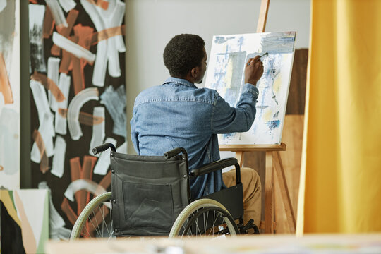 Rear view of African American male painter in wheelchair painting on canvas while sitting in front of easel in studio or classroom