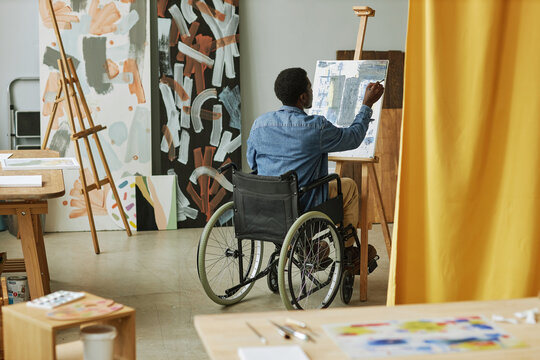 Back view of young black man sitting in wheelchair in front of easel and painting on canvas with acrylic paints in studio of arts