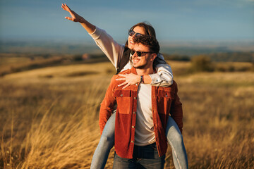 A boy is carrying a girl on his back, while she is showing him something in the distance. The couple wears sunglasses and seems happy.